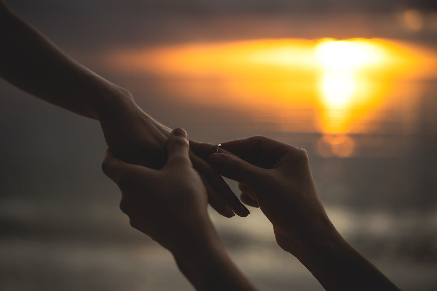 海に沈む夕日を背景に新婦の薬指へ指輪をはめる新郎の手元