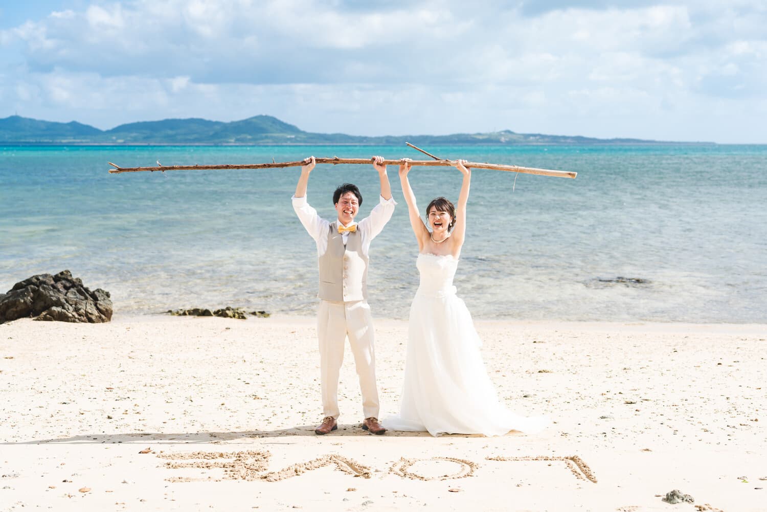 砂浜に文字を書いて流木を掲げる新郎新婦