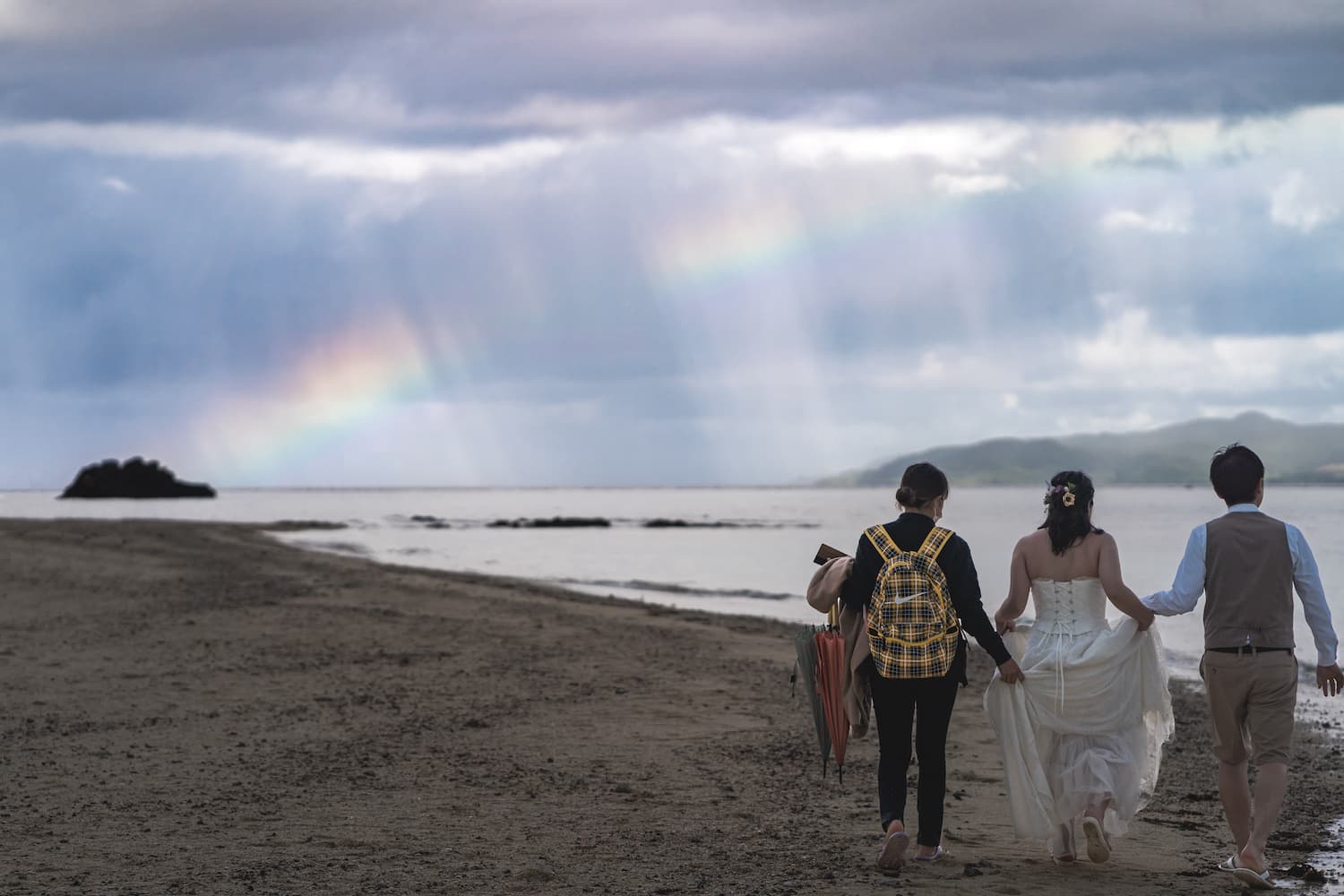 海に架かる虹を背景に浜辺を歩いている新郎新婦と撮影スタッフ